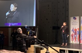 Nainen puhumassa esiintymislavalla vieressä iso screeni ja edessä tv-kamera. 