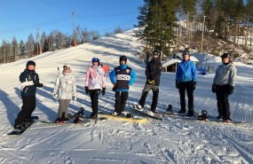 Seitsemän laskettelijaa vierekkäin poseeraamassa taustallaan Hyvinkään Sveitsin talvinen ja aurinkoinen rinne.