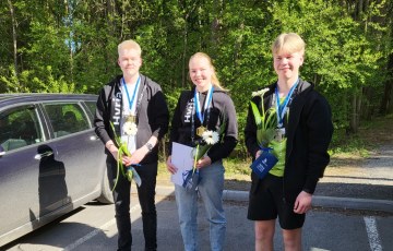 Kolme hymyilevää nuorta opiskelijaa seisomassa vierekkäin parkkipaikalla.