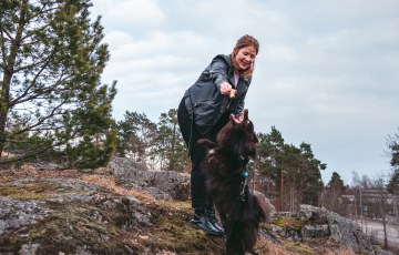 Nainen ja koira leikkimässä kalliolla. Taustalla mäntyjä ja taivasta. 