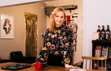 Nainen seisoo pöydän takana ja nojaa siihen kodinomaisesti sisustetulla toimistolla